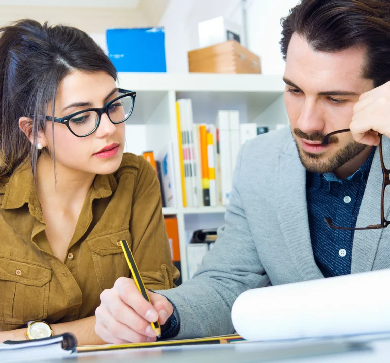 Two individuals engaged in a collaborative discussion at a workspace, with one writing notes and the other holding glasses,